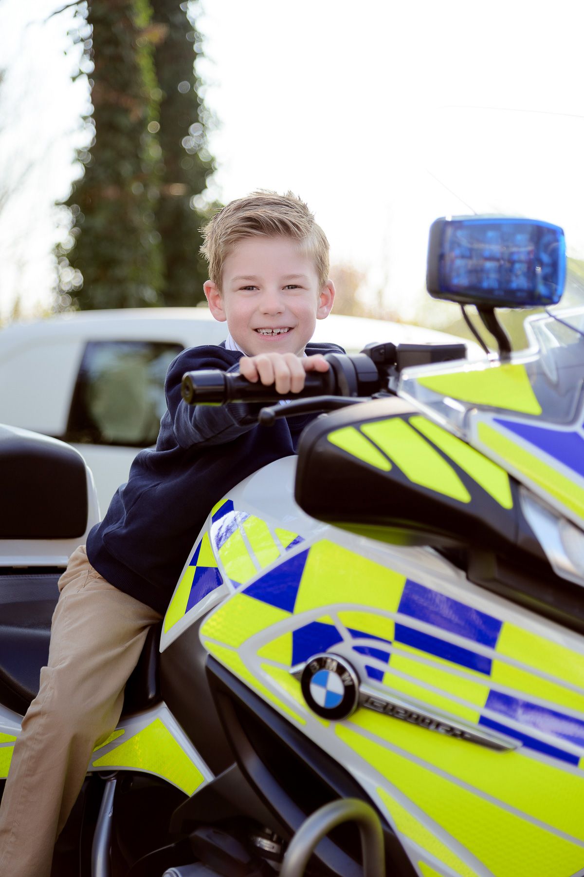 Young boy smiling on a BMW police motorcycle, wearing a navy top and khaki pants outdoors.