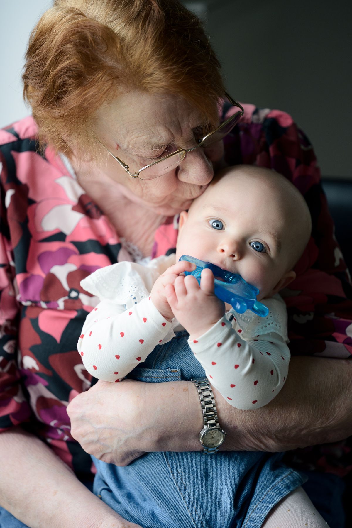 An elderly grandmother kisses a blue-eyed baby holding a toy while seated indoors.