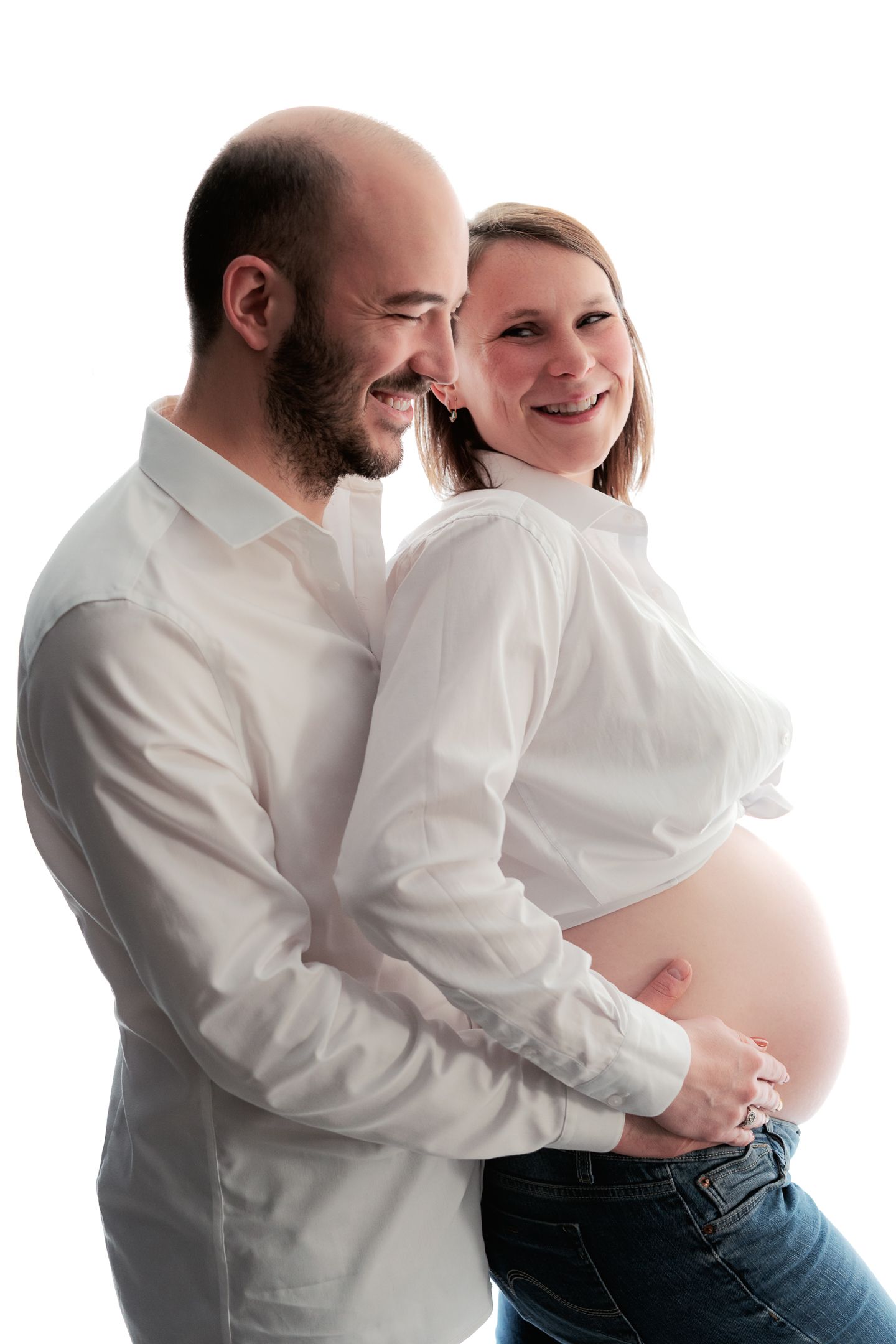 Happy couple in white shirts, pregnant woman with exposed baby bump, man embracing her from behind, studio photo.