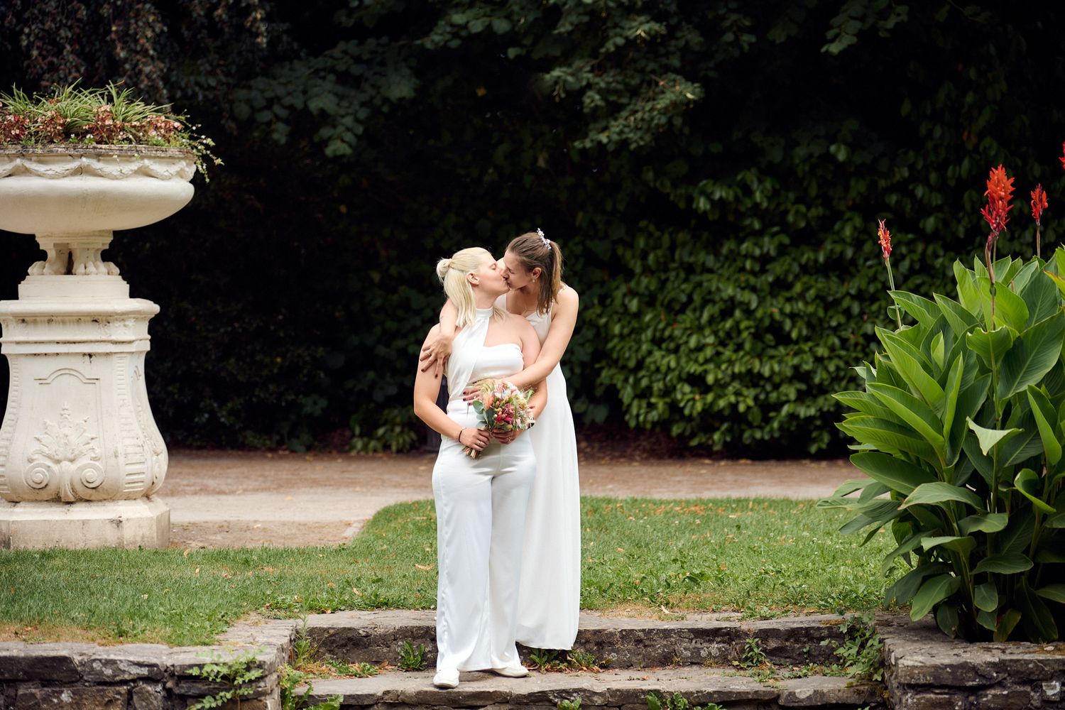 Two brides in white embrace outdoors near a stone urn with flowers, surrounded by lush greenery.