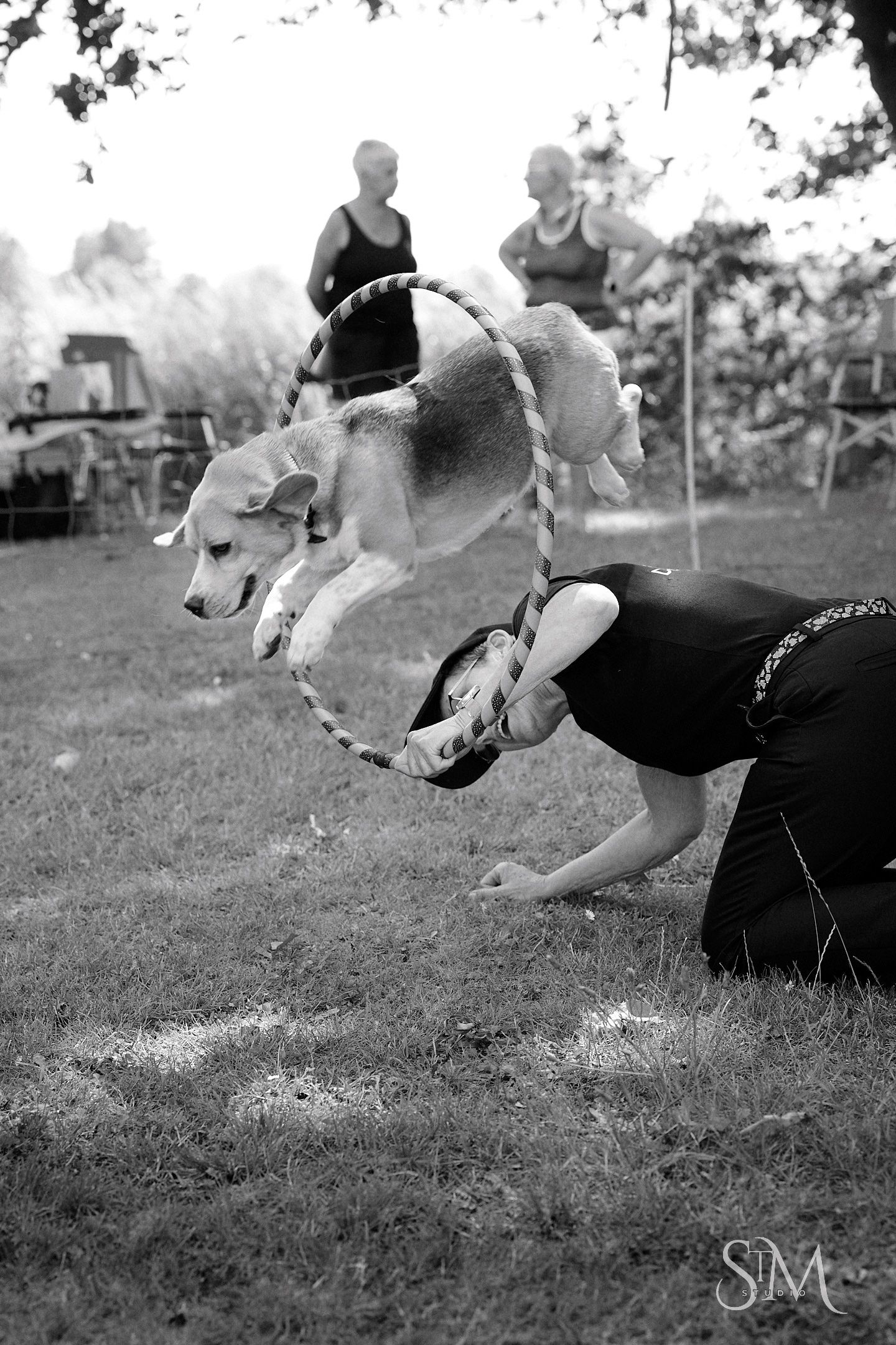 Black and white photo of a dog leaping through a hoop held by a kneeling trainer at an outdoor dog agility event.