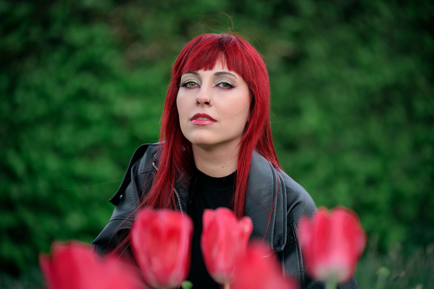Woman with bright red hair posing outdoors near red tulips with a green hedge background.