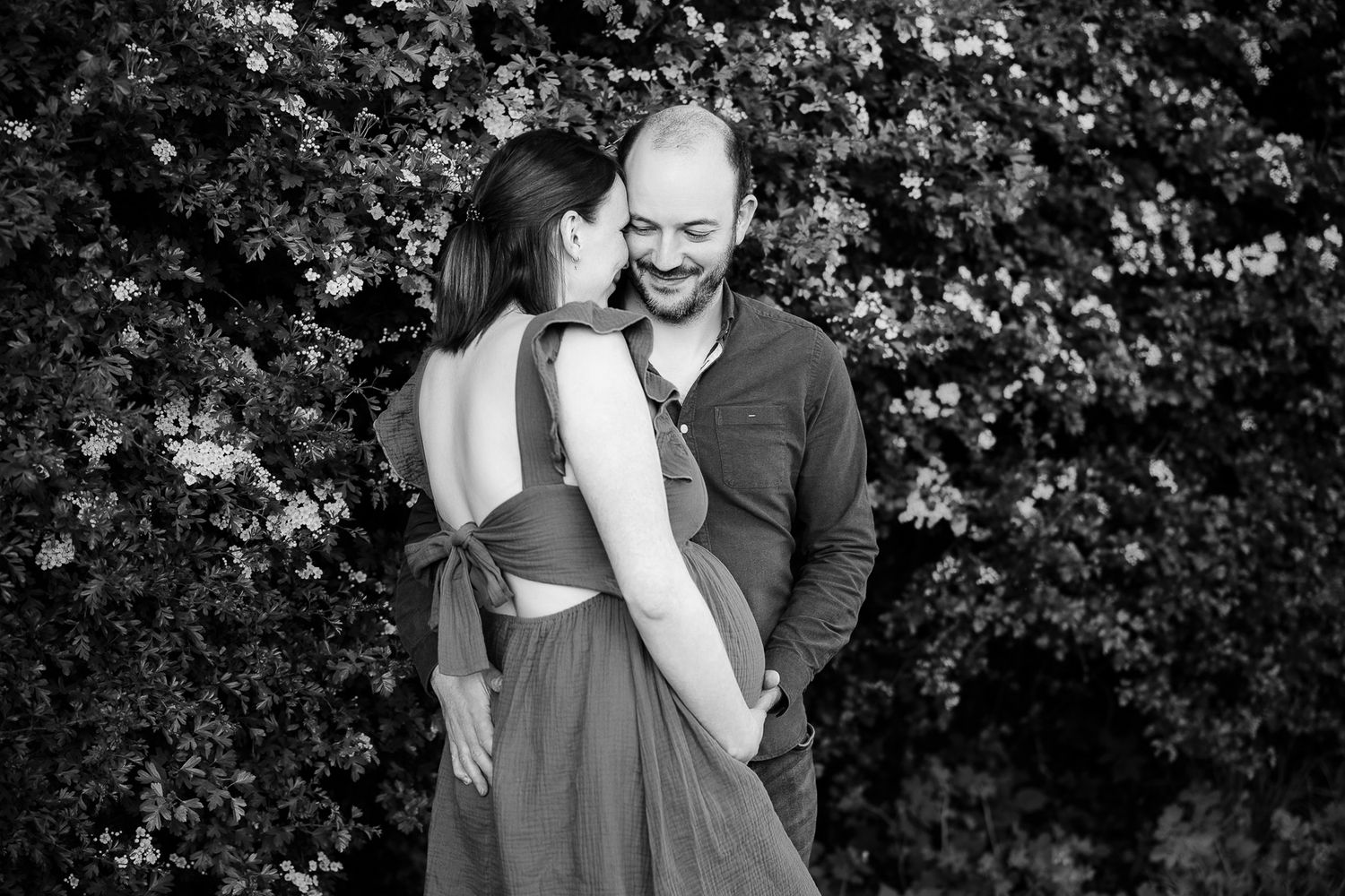 Black and white photo of a smiling couple embracing in front of lush flowering bushes outdoors.
