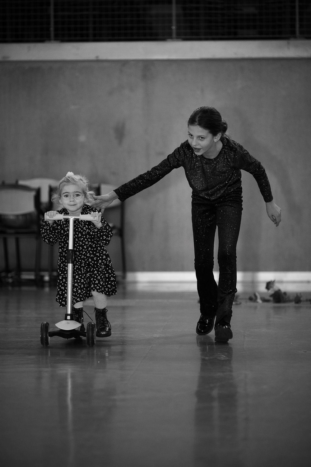 Black and white photo of older girl helping young child ride a scooter on an indoor skating rink.