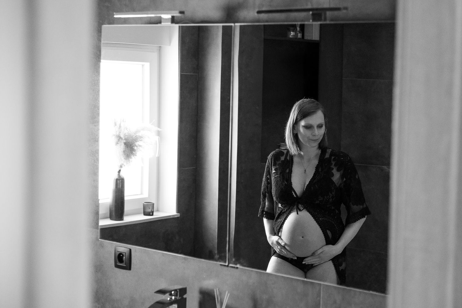 Black and white photo of a pregnant woman in lingerie holding her baby bump, reflected in a bathroom mirror.