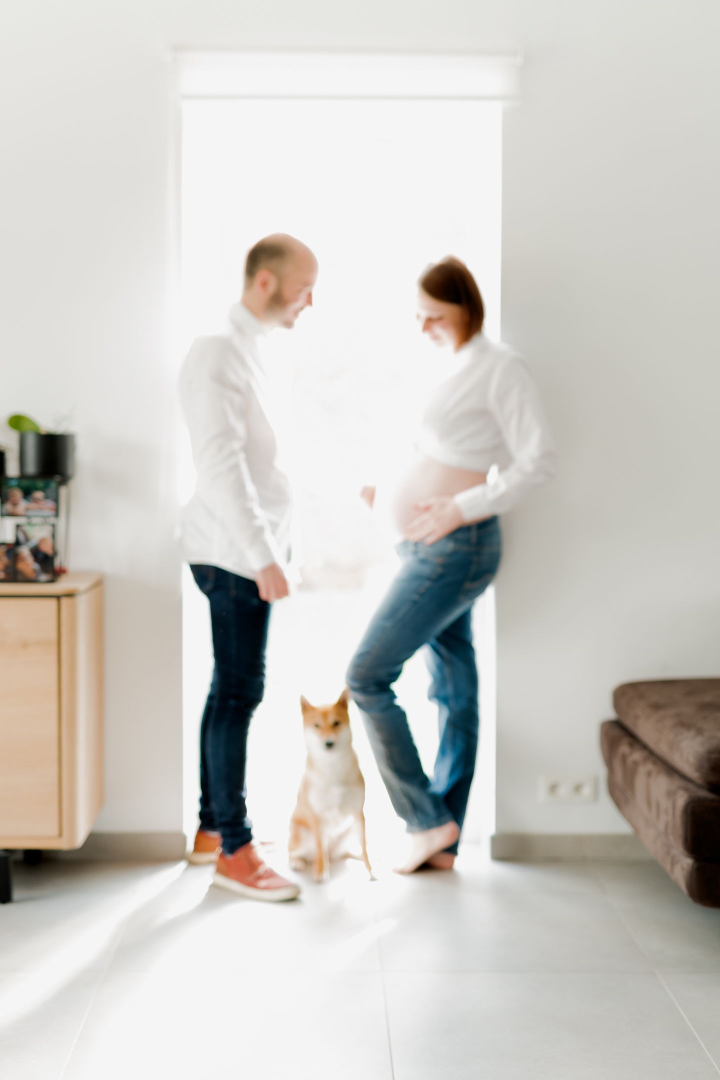 Pregnant woman and man in white shirts stand with a Shiba Inu dog in a bright modern living room.