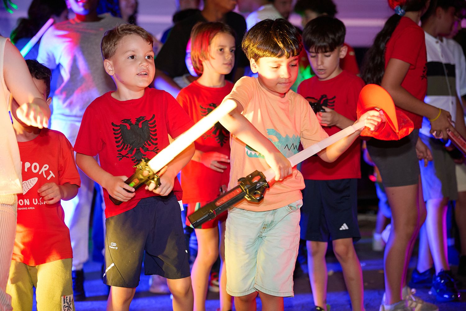 Children playing with toy swords at a colorful outdoor event, wearing red shirts and casual summer clothing.