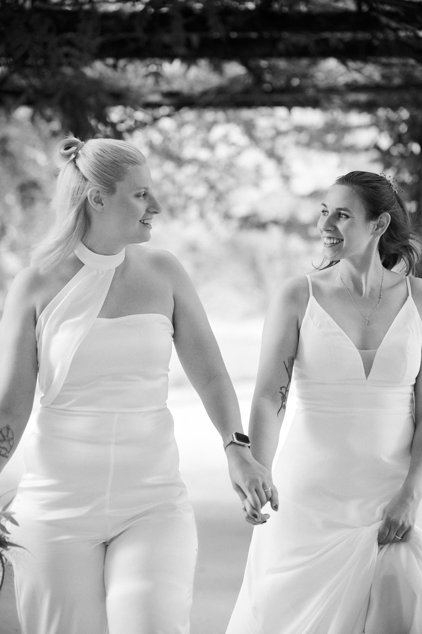 Two brides in white gowns hold hands and smile at each other in a romantic black and white wedding photo.