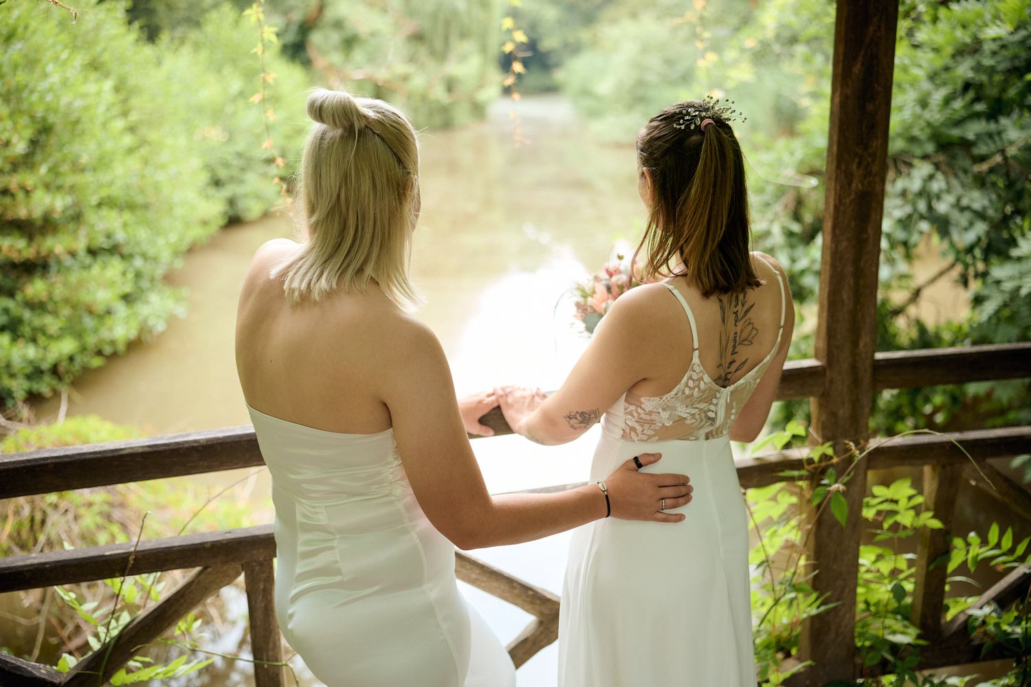 Two brides in white dresses stand on a wooden deck overlooking a serene river surrounded by lush greenery.