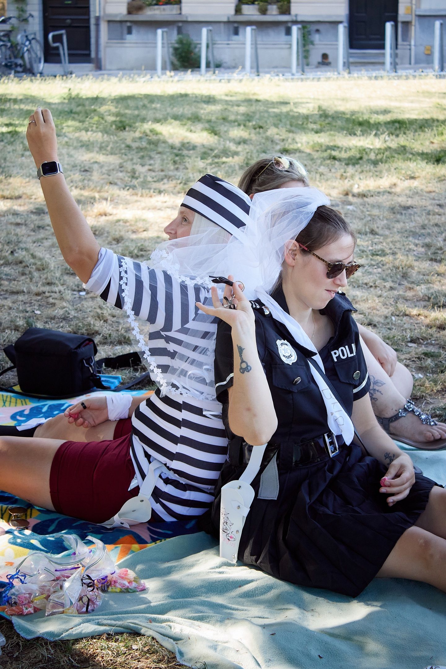 Two women in costume outfits, one as a prisoner and one as a police officer, sitting on grass at an outdoor event.