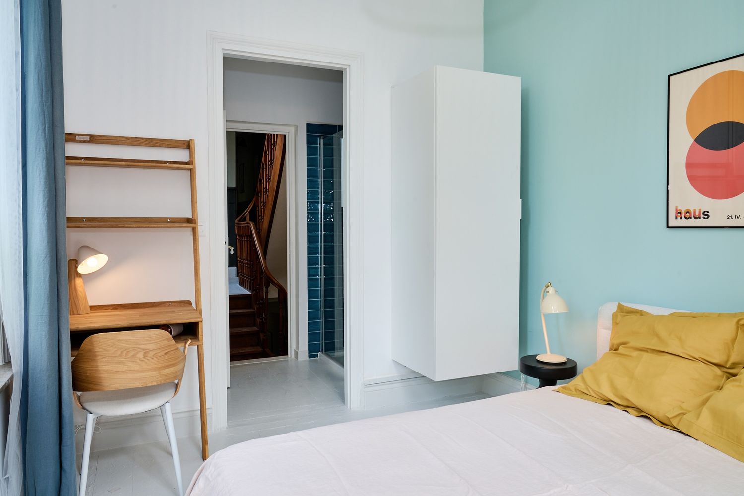 Modern bedroom with teal walls, yellow bedding, white wardrobe, wooden desk, and en-suite bathroom glimpse.
