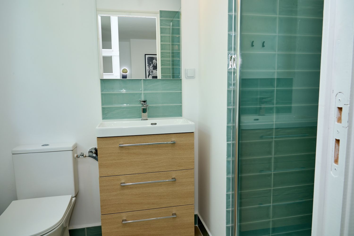 Modern bathroom with green tiles, wood vanity, mirror, toilet, and glass shower enclosure.