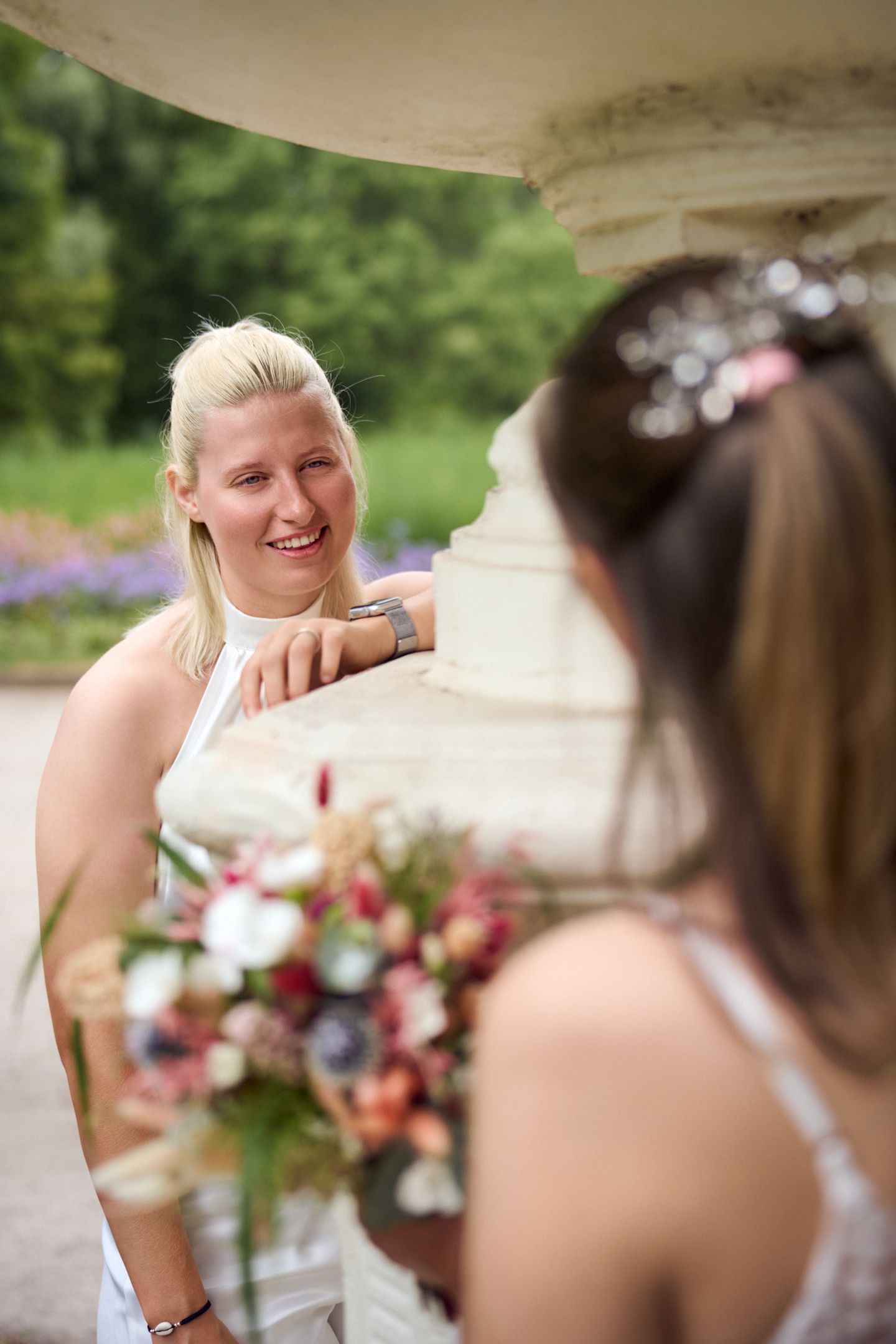 Two brides in white dresses laughing together outdoors, holding colorful wedding bouquets near stone columns.