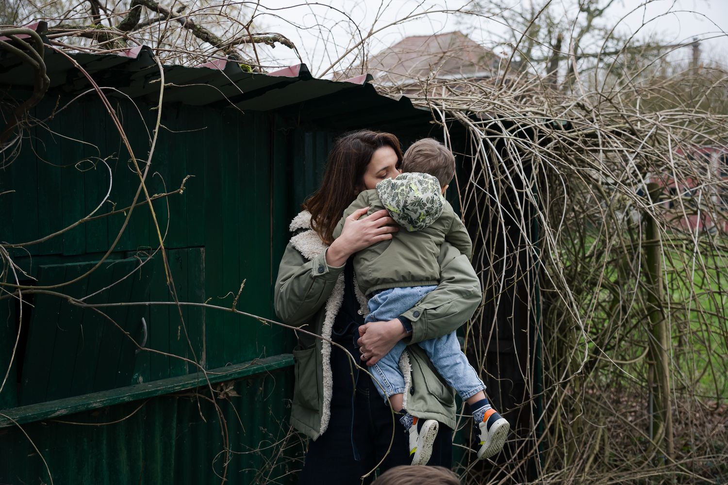 Mother in a green jacket holding and embracing her young child outdoors near a fence.