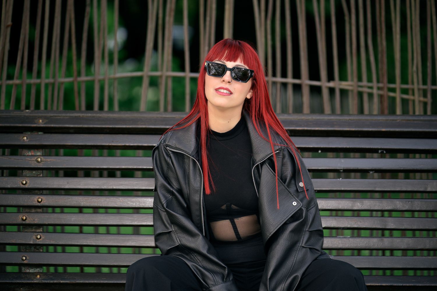 Woman with red hair in black leather jacket and sunglasses sitting on park bench.