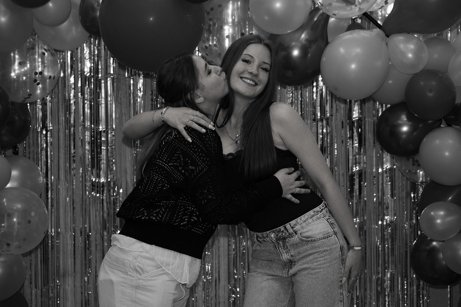 Two women posing together at a party with balloon arch and silver tinsel backdrop in black and white photo.