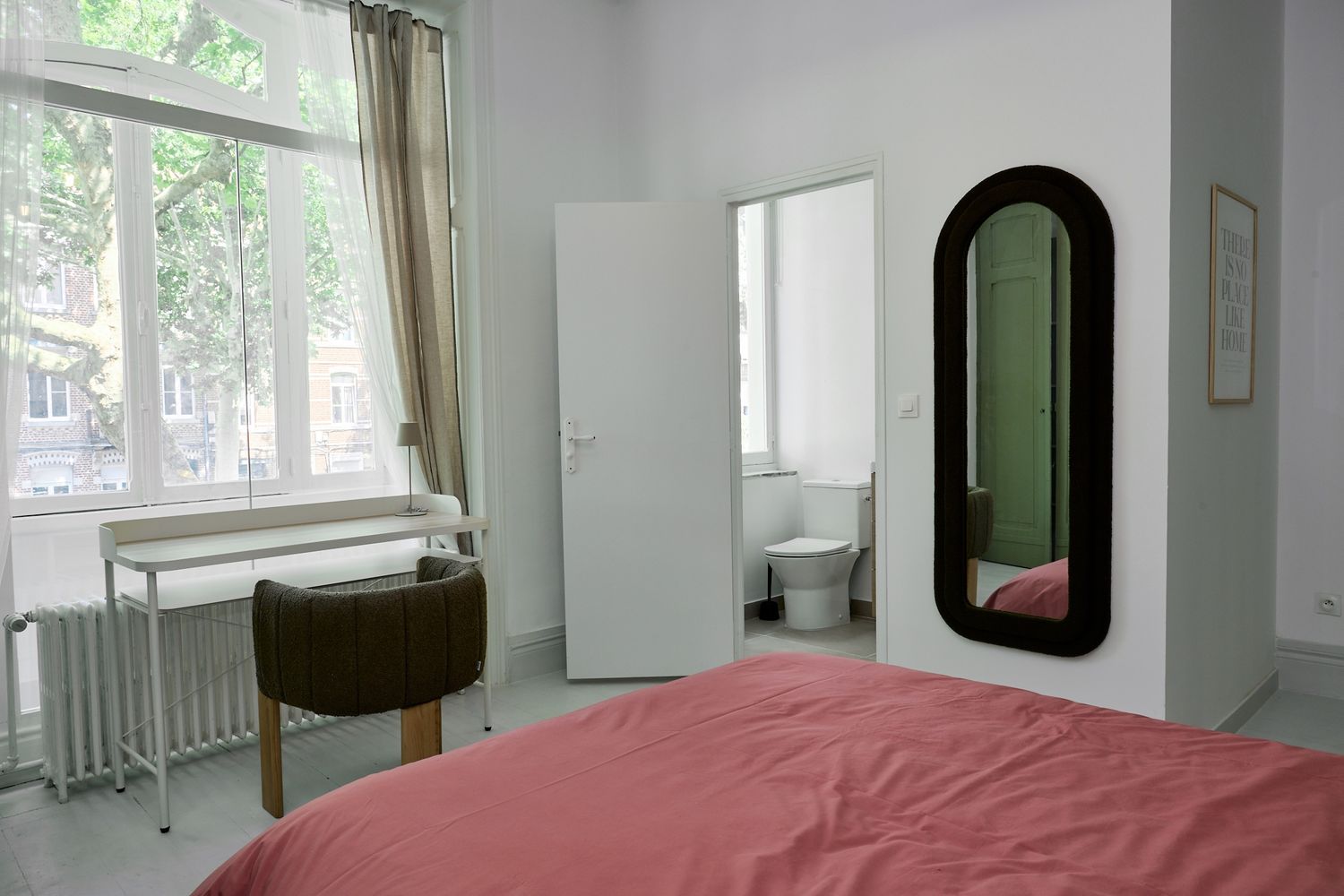Bright minimalist bedroom with pink bedding, arched mirror, white wardrobe, and large window overlooking trees.