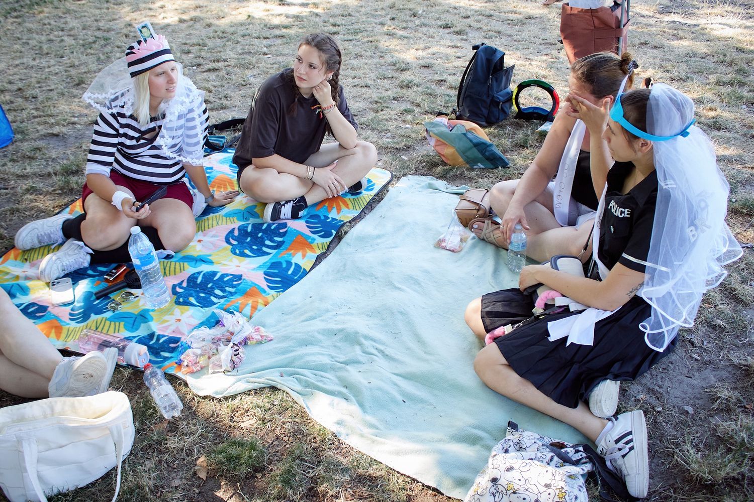 Group of young people sitting on colorful blanket in park, playing cards and relaxing outdoors on sunny day.