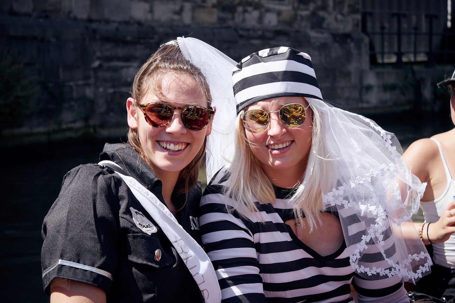 Two women in festive costumes with sunglasses smile outdoors, one wearing a striped outfit and bridal veil.