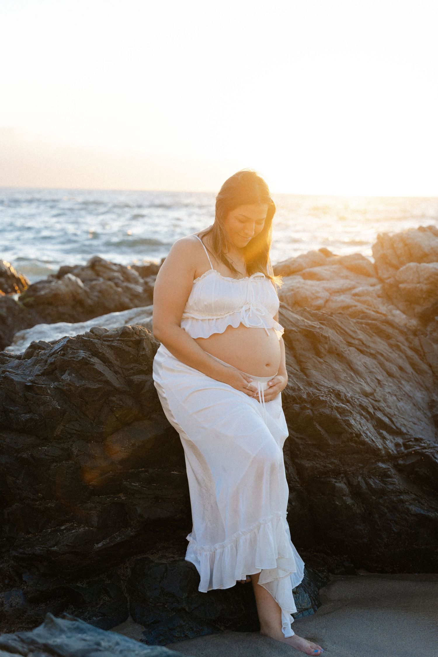 Pregnant woman cradling her baby bump on a rocky beach at golden sunset Conchas chinas beach - Marina Pochepkina photography