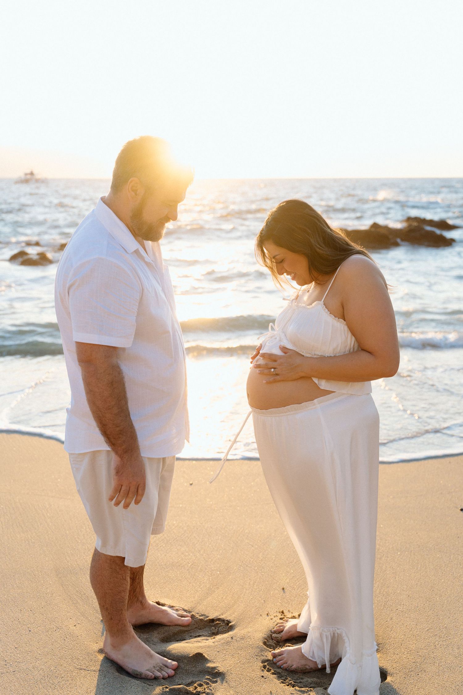 Couple in white outfits on a sunset beach, man gazing at pregnant woman cradling her baby bump in Punta Mita, Riviera Nayarit