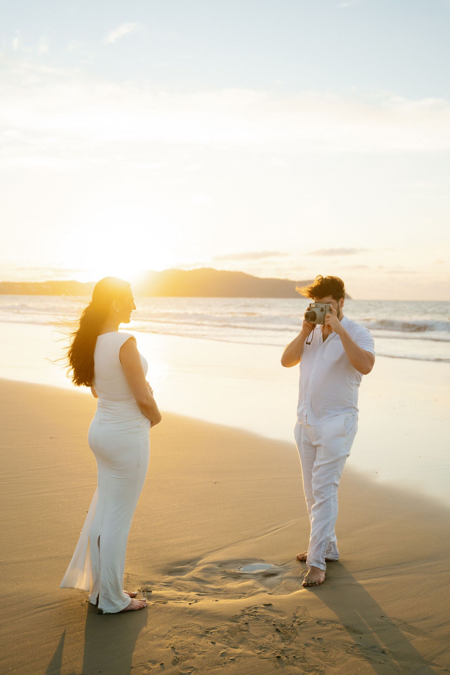 Couple on sandy beach at golden sunset, man photographing woman by ocean waves Conrad hotel - Marina Pochepkina photographer