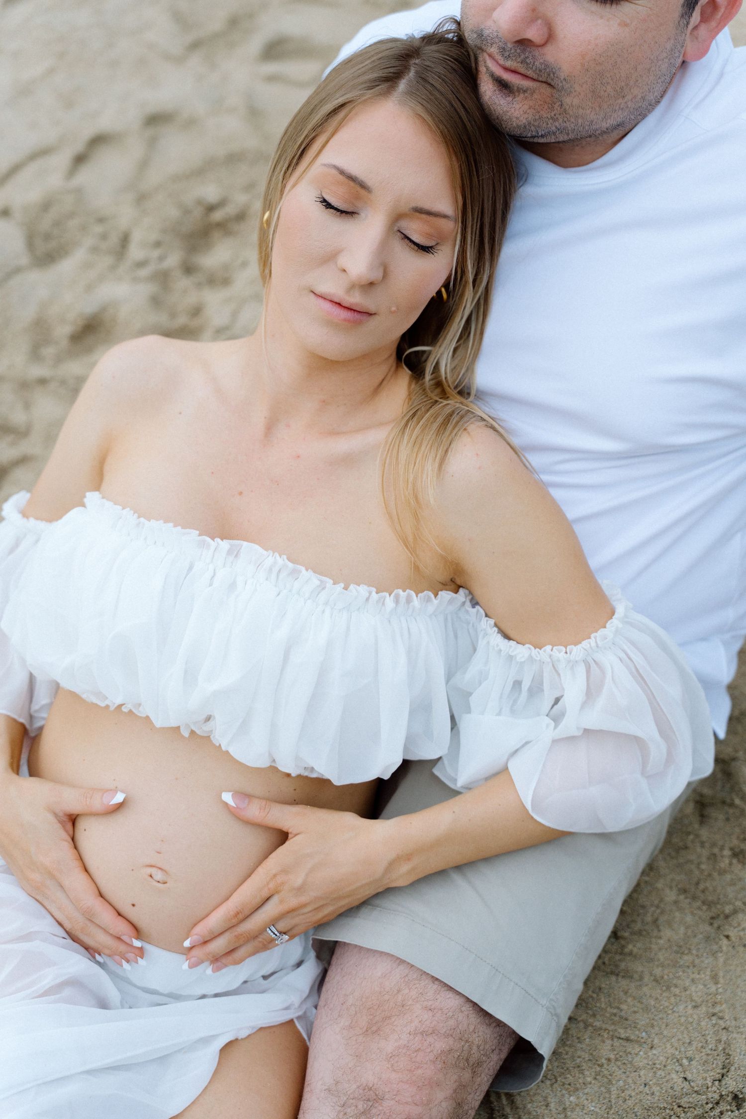 Pregnant woman sits on sand, cradled by partner in white, both tenderly touching her baby bump in in Puerto Vallarta — Marina Pochepkina