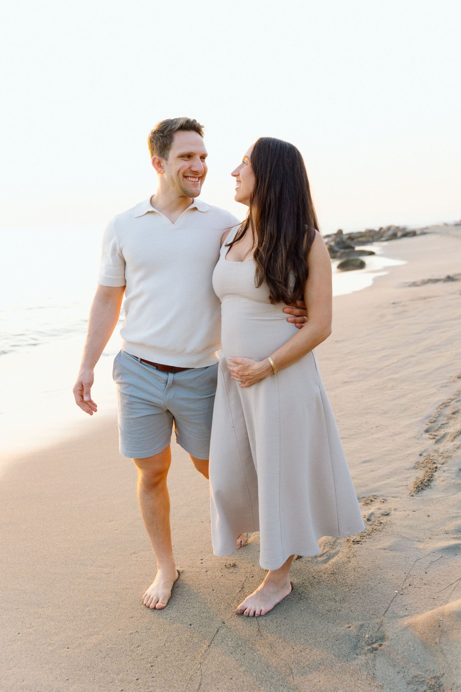 A smiling couple walks barefoot on a sandy beach at sunset, the woman cradling her baby bump in Puerto Vallarta- Marina Pochepkina photo