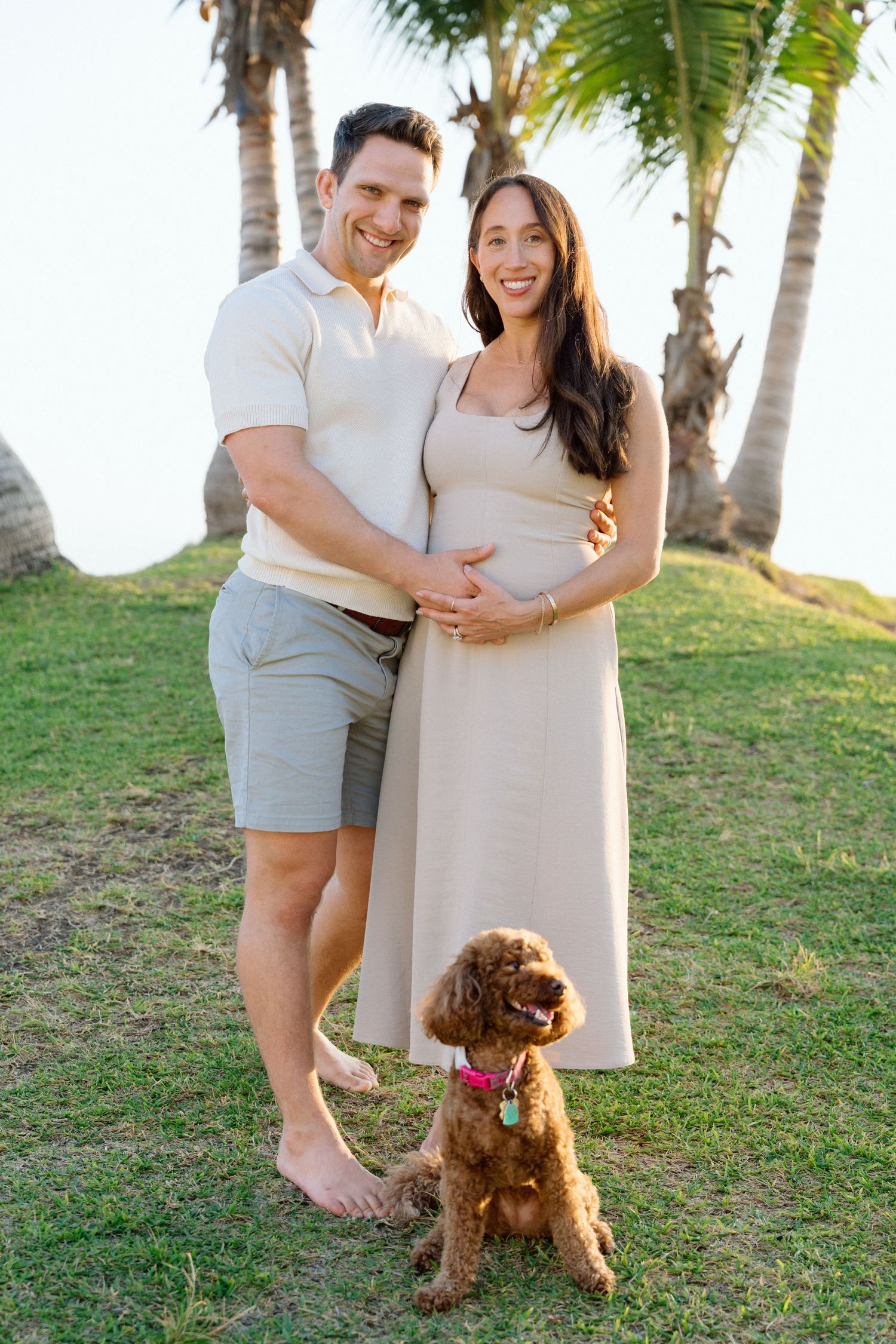 Smiling couple poses outdoors near palm trees, holding baby bump, with a brown doodle dog in Puerto Vallarta - Marina Pochepkina photography