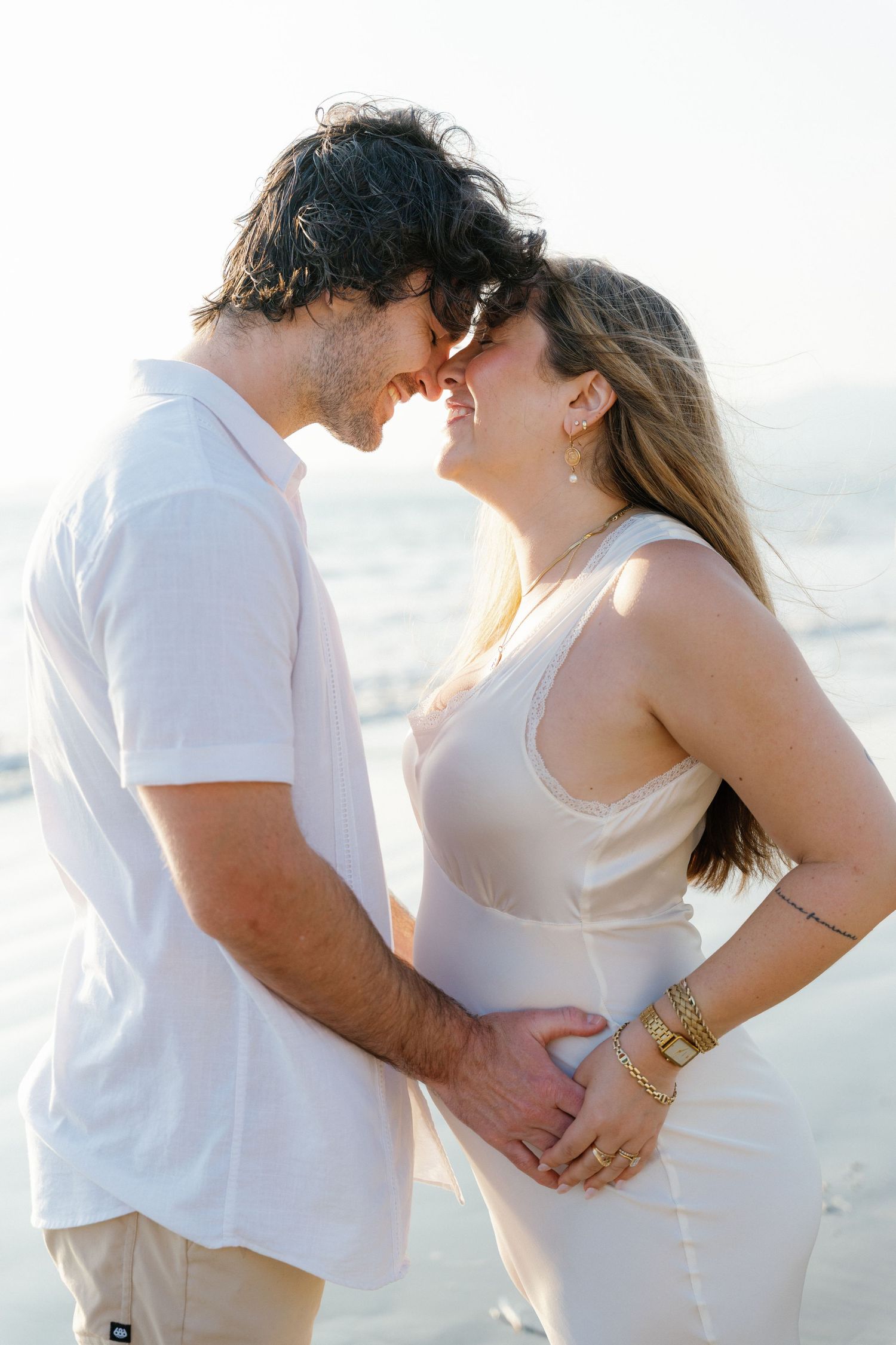 A couple in white outfits share a romantic moment on a sunlit beach, touching foreheads and smiling in Puerto Vallarta -Marina Pochepkina