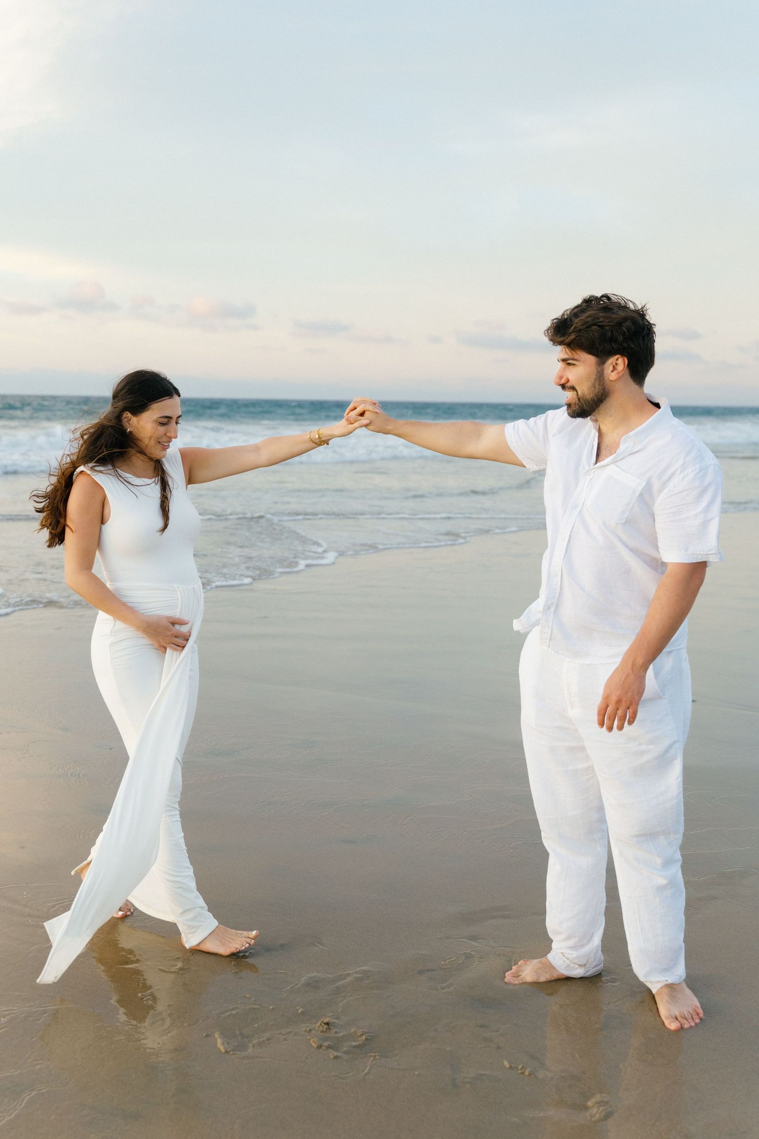 Pregnant couple holding hands on a beach at sunset, barefoot on wet sand with ocean waves behind them in Punta Mita - Marina Pochepkina
