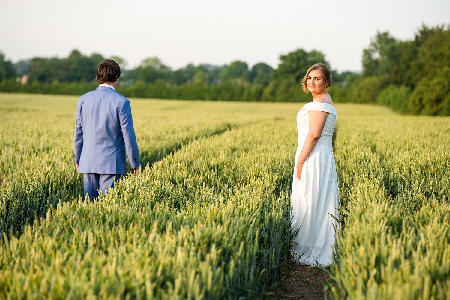 Bride in white gown and groom in blue suit holding hands in a lush green wheat field at golden hour.