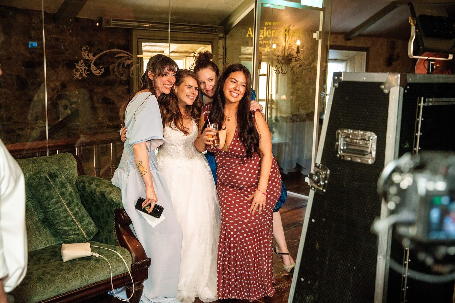 Three smiling women in a white dress and polka dot dresses pose together near a photo booth and green sofa at a festive indoor party.