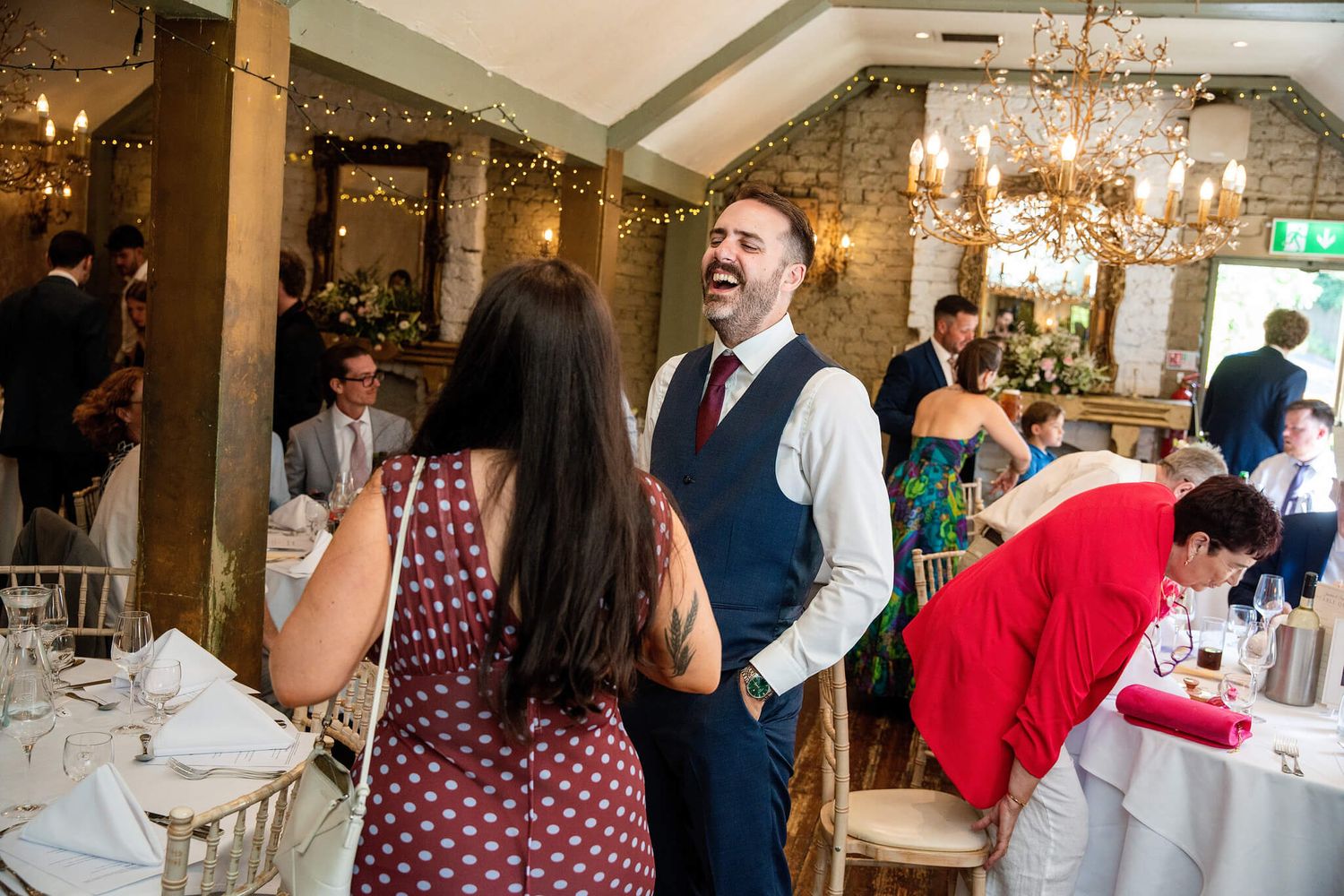 Smiling man in a vest and tie laughs with a woman at a festive wedding reception with chandeliers, stone walls and set tables.