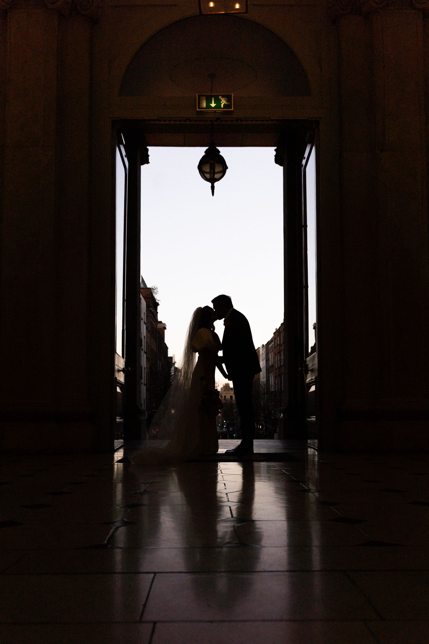 Silhouette of bride and groom kissing in arched doorway with soft backlight, romantic wedding portrait.