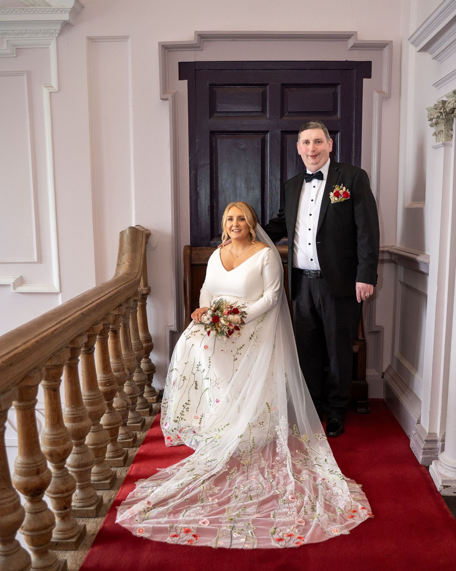 Bride in white gown with floral veil and groom in tuxedo pose on red-carpeted staircase in elegant venue at Bellinter house.