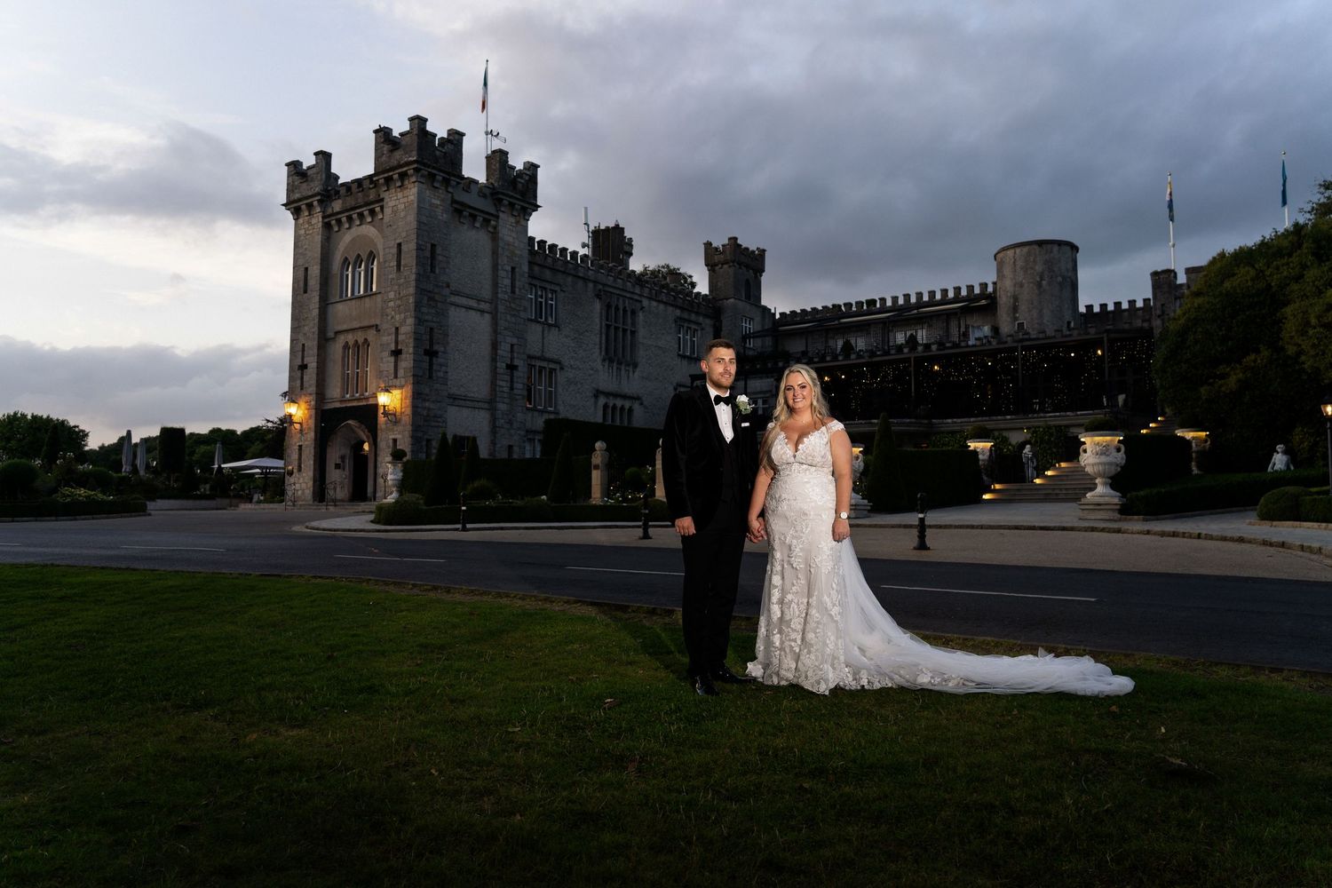 Two people in wedding attire pose at dusk in front of a majestic stone castle with dramatic cloudy skies.
