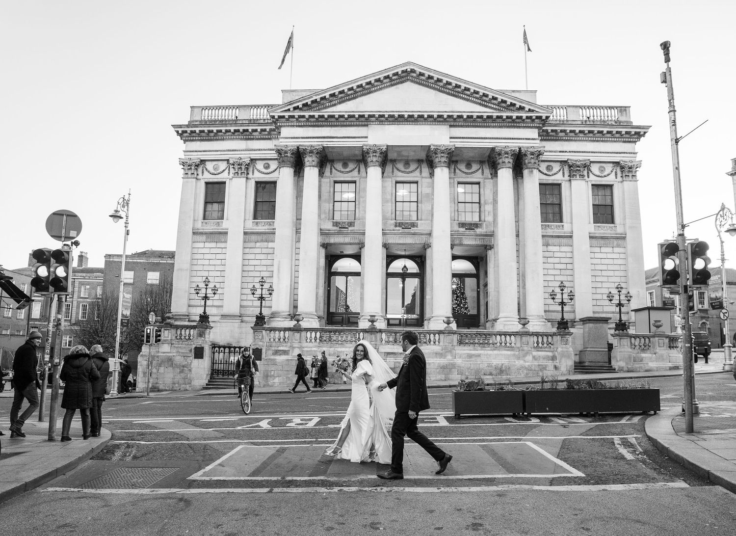 A panoramic black and white view of a neoclassical building with columns and people walking in front.