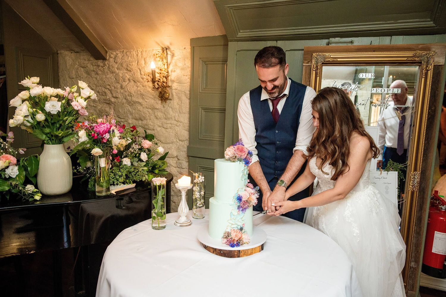 Bride in a white gown and groom in a navy vest cutting a pastel floral wedding cake together beside fresh flower arrangements.