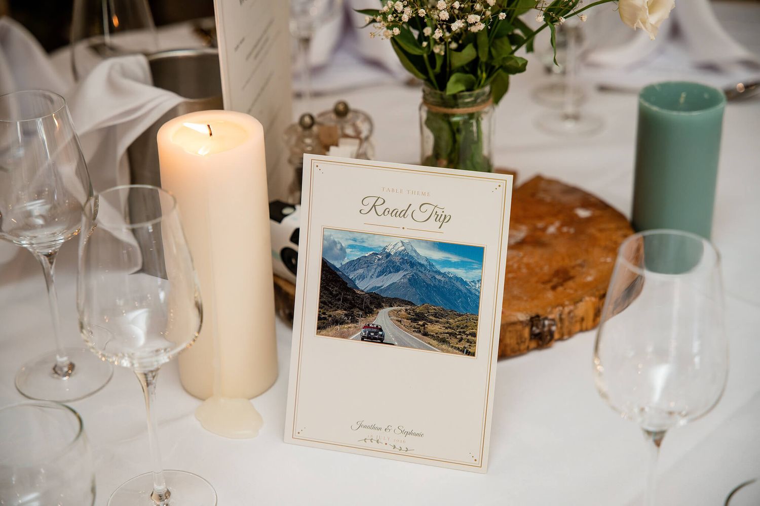 Wedding reception table with a Road Trip themed card showing a mountain landscape, beside a lit pillar candle, flowers and wine glasses.