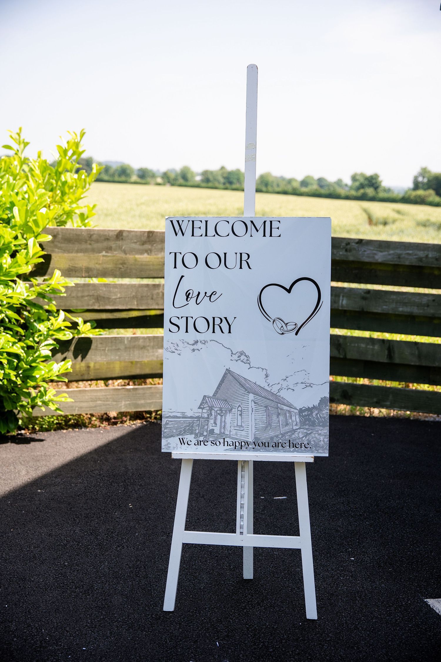 White easel wedding welcome sign reading Welcome To Our Love Story outdoors at a rustic venue.
