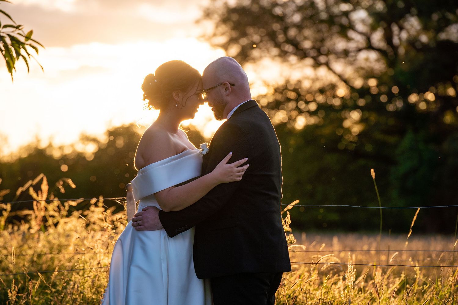 Bride and groom embracing at golden sunset in a field, romantic outdoor wedding portrait.