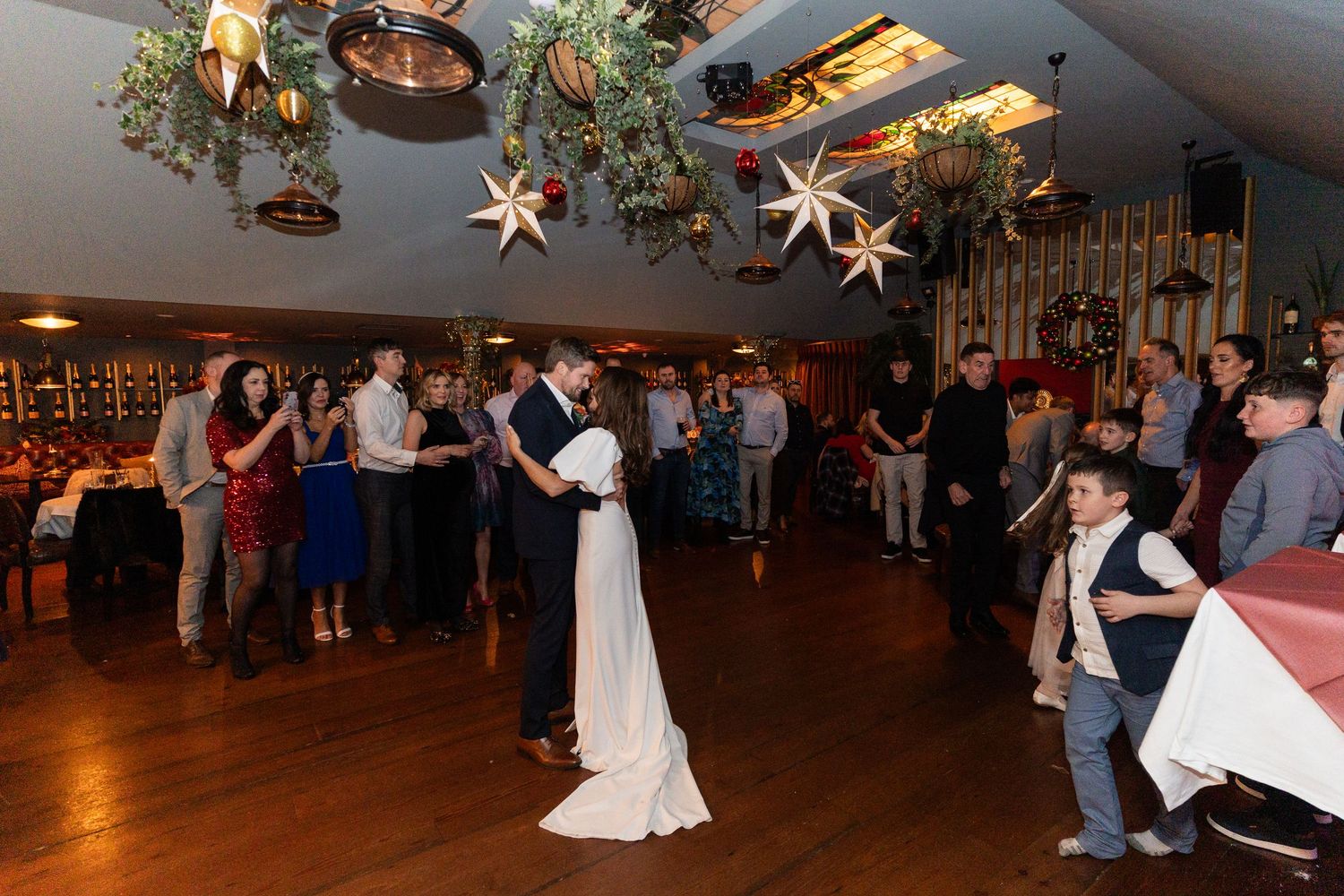 Bride and groom share their first dance surrounded by guests in a festively decorated wedding reception venue.