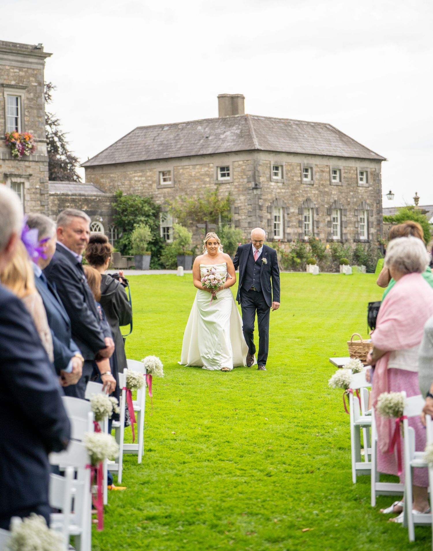 Bride in a white gown walks down a grass aisle with an older man toward seated wedding guests, with a historic stone manor behind them.