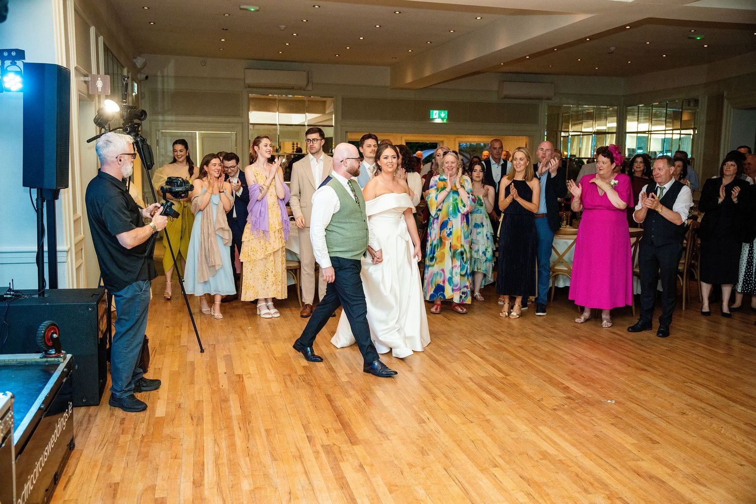 Bride and groom enter wedding reception on dance floor surrounded by applauding guests in elegant venue.