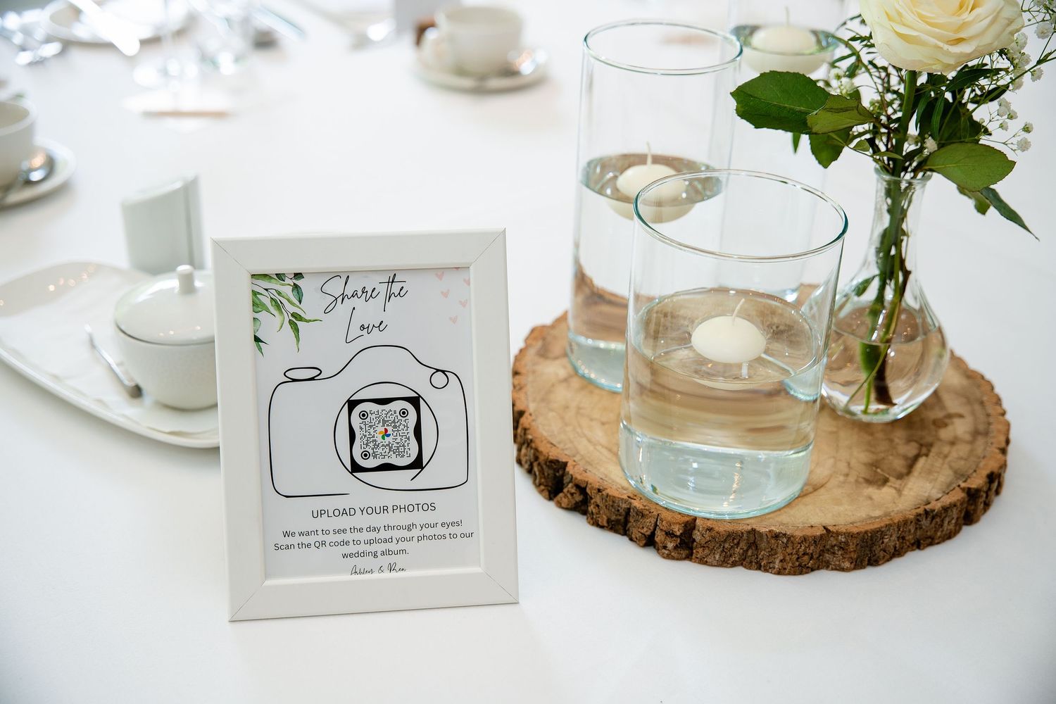 Wedding table decor with QR code photo-sharing sign, glasses of water, white roses, and a rustic wood slice centerpiece.