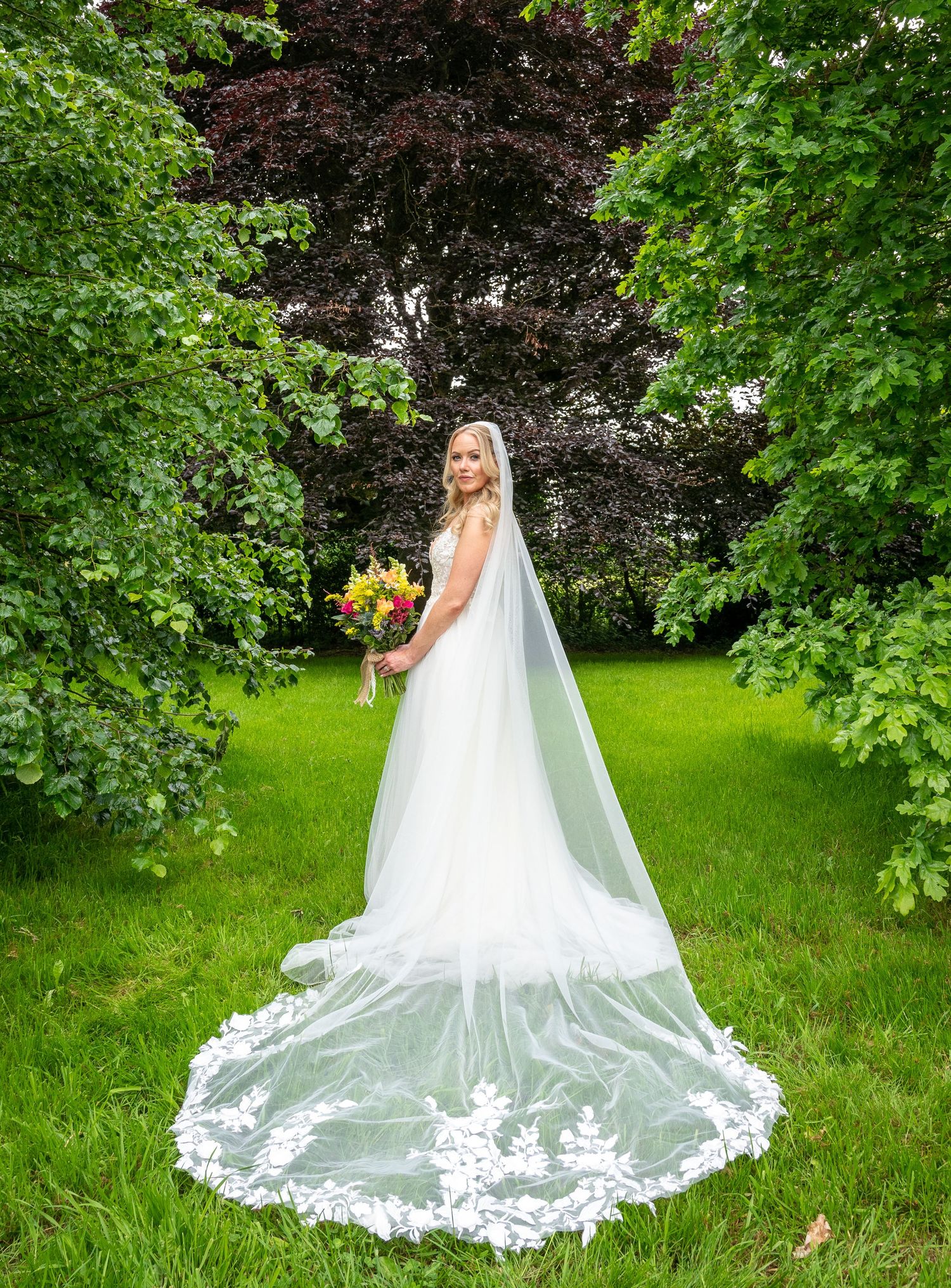 Bride in white gown with lace cathedral veil holding colorful bouquet standing in lush green garden at Bellinter house.