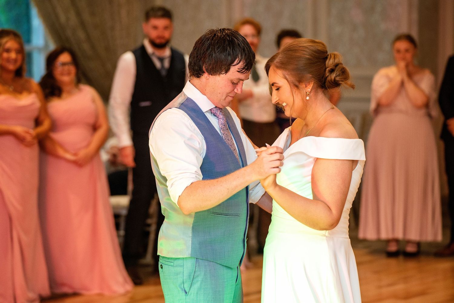 Bride and groom share their first dance at wedding reception, surrounded by guests in pink bridesmaid dresses.