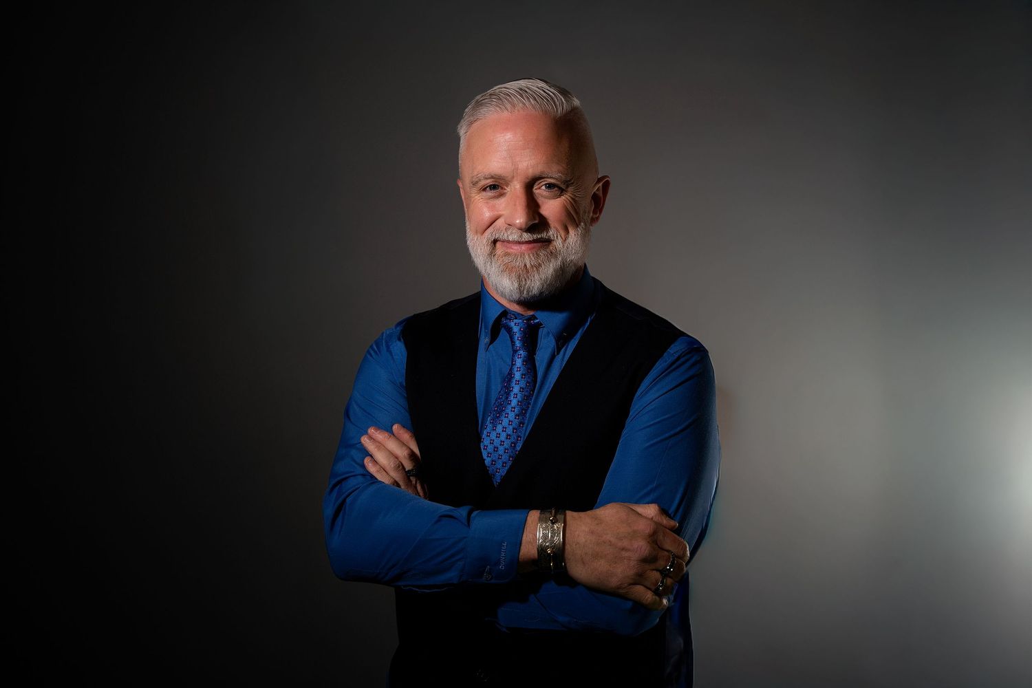 Professional man with gray beard, crossed arms, wearing blue shirt and vest, posing confidently against dark background.