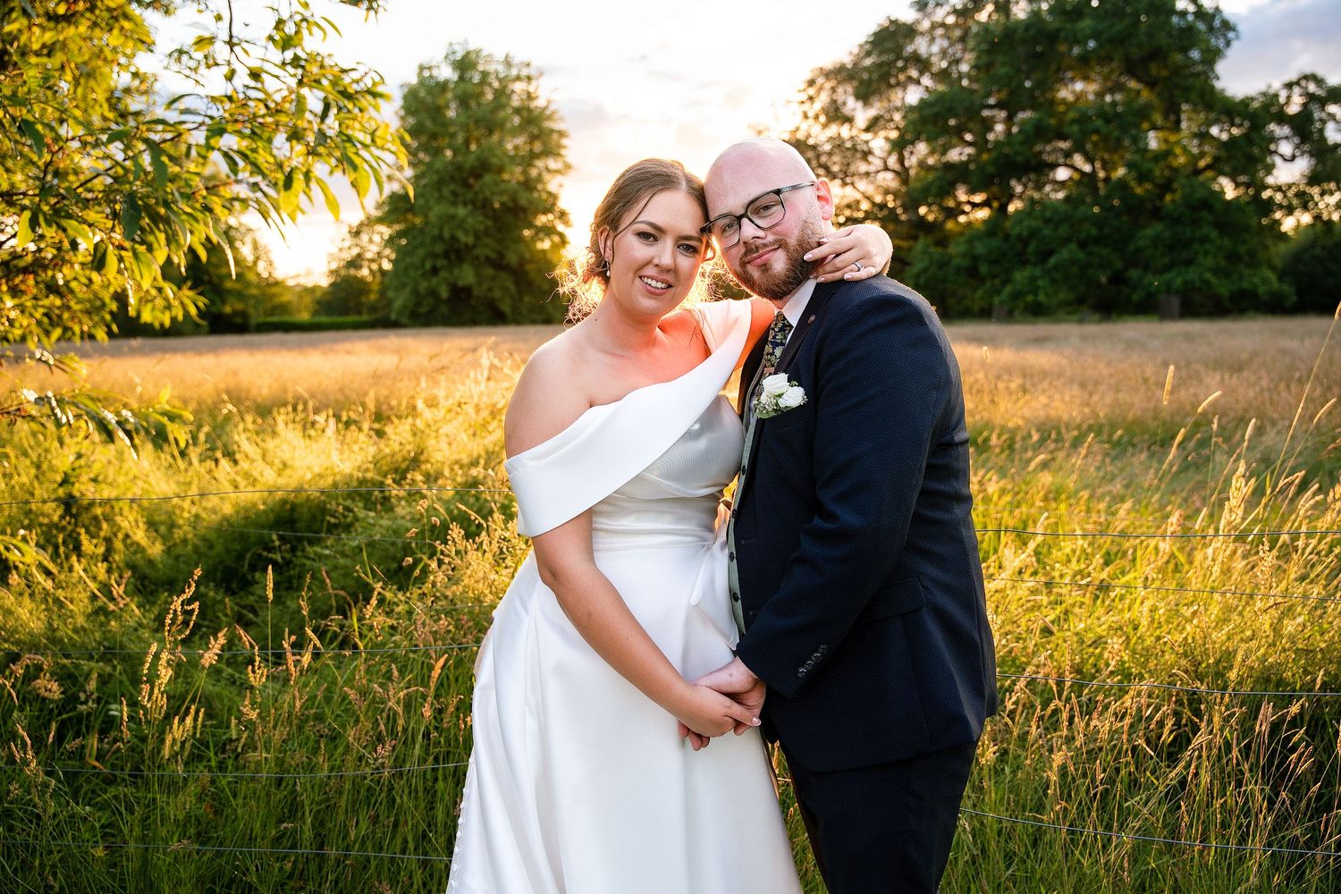Bride and groom embracing in golden hour sunlight in a lush green field, bride in white off-shoulder gown.